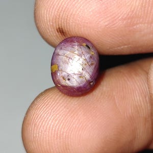 May include: A close-up of a polished, oval gemstone. The gem is a deep reddish-purple colour with internal golden inclusions and a few small yellow spots. The gem is held between two fingers.