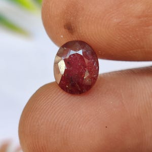 Puede incluir: Una gema facetada de forma ovalada con un tono rojo intenso. La gema se sostiene entre dos dedos, mostrando su superficie pulida y claridad interna. La piedra, probablemente un rubí, refleja la luz con un brillo sutil.