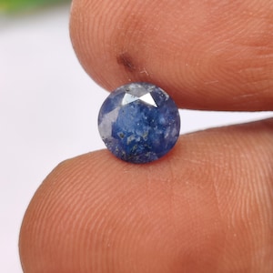 Puede incluir: Una gema de zafiro redonda y facetada. El zafiro es de color azul profundo con inclusiones azules claras y blancas. La gema se sostiene entre dos dedos, mostrando su tamaño y detalle. El corte de la piedra crea múltiples facetas.
