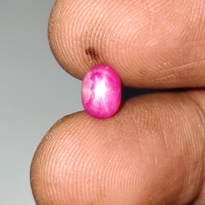 May include: A small, oval-shaped, cabochon-cut gemstone with a vibrant magenta-pink colour. The stone displays a distinct six-rayed star effect, known as asterism, visible on its polished, smooth, and rounded surface under direct light.