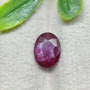 May include: An oval-shaped, faceted ruby gemstone with a deep magenta-red color and a textured, crystalline surface. The stone features a polished, multi-faceted top that reflects light, showcasing its natural inclusions and opaque, rich red hue.