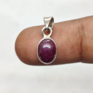 Könnte beinhalten: Ein kleiner, ovaler Anhänger mit einem tiefroten Edelstein in einem silbernen Metallrahmen. Der polierte Edelstein hat eine satte rote Farbe. Der Anhänger hat oben eine Schlaufe für eine Kette. Der Anhänger wird von einem Finger gehalten.