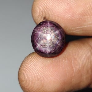 Könnte beinhalten: Ein runder, im Cabochon-Schliff gehaltener Sternrubin-Edelstein mit einem tiefen purpurroten Farbton. Der Stein weist einen ausgeprägten sechsstrahligen Sterneffekt auf, bekannt als Asterismus, der als helles, reflektierendes Muster erscheint.