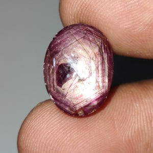 May include: An oval-shaped gemstone with a star-like pattern in shades of pink and white. The gemstone is set in a silver ring.