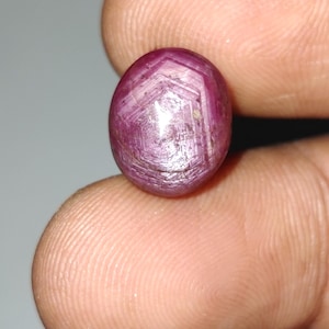 May include: Oval-shaped, deep red ruby gemstone. The polished surface reflects light, revealing internal hexagonal patterns. The gemstone is held between fingers, showcasing its size and color.