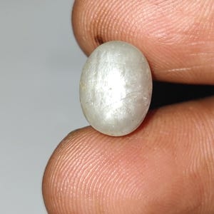 May include: A pale, oval-shaped gemstone, held between fingers, exhibits a soft, pearly luster. The stone's surface has subtle internal lines, giving it a unique texture. The overall color is a creamy white, with hints of iridescence.