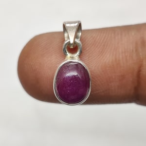 Pode incluir: Um pingente de prata com uma pedra preciosa ovalada de cor vermelha escura. A pedra é engastada em uma moldura de prata e tem uma superfície polida e brilhante. O pingente tem um design simples e elegante, adequado para uso diário.