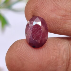 Puede incluir: Una piedra preciosa ovalada de color rojo oscuro con un corte facetado. La piedra preciosa se sostiene en una mano.