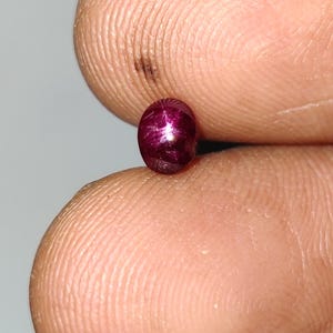 Puede incluir: Una pequeña piedra preciosa de rubí estrella con corte cabujón ovalado y un color rojo magenta profundo y saturado. La piedra presenta un efecto de asterismo distintivo de seis rayos, donde la luz se refleja en un patrón de estrella.