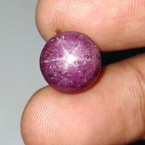 May include: A spherical, polished, purplish-red gemstone displaying a six-rayed star effect. The gem is held between fingers, highlighting its colour and the star pattern. The gem is approximately 1.9 cm in diameter.