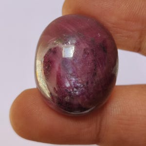 May include: A polished, oval-shaped gemstone with a deep burgundy hue. The stone has a translucent quality, revealing internal patterns and inclusions. The surface reflects light, creating highlights and shadows. The gemstone is held between fingers.
