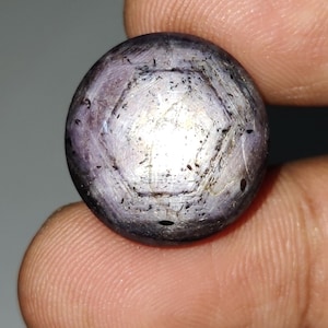 May include: A spherical, polished gemstone in a deep purple shade. The gem displays a six-sided design on its surface, with a lighter, metallic-like sheen at the centre. The stone is held between two fingers, highlighting its size and form.