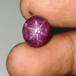 Op de afbeelding: Een ronde, cabochon geslepen sterrobijn edelsteen met een diepe magenta-rode kleur. De steen vertoont een duidelijk zesstralig asterisme-effect, dat verschijnt als een helder wit sterpatroon over het gepolijste, doorschijnende oppervlak.