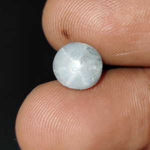 May include: A round, pale blue gemstone with a star-like pattern. The gem is held between two fingers against a dark background. The stone's surface reflects light, creating a subtle shimmer.