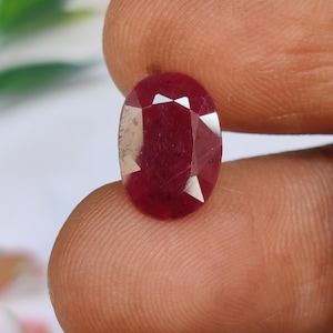 May include: A faceted, oval-shaped ruby gemstone. The gem is a deep red color with multiple facets that reflect light. The ruby is held between two fingers, showcasing its size and shape.