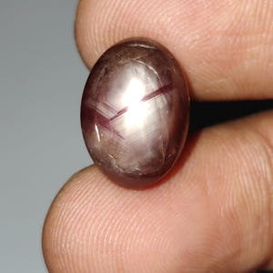 Puede incluir: Gema ovalada, tallada en cabujón, con efecto estrella. La gema es de color marrón púrpura oscuro con un patrón de estrella de seis rayos. La piedra es lisa y pulida, reflejando la luz.