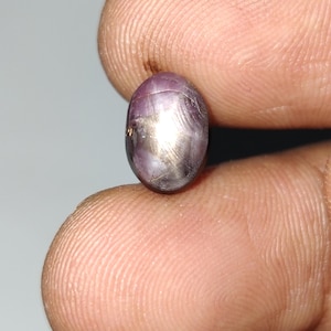 May include: A small, oval, purplish-red gemstone with a star effect. The gem is held between fingers, highlighting its polished surface and natural colour variations. The stone appears to be a star ruby.
