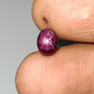 May include: A small, oval-shaped, deep magenta-colored gemstone with a smooth, polished surface. The stone displays a distinct six-rayed star pattern across its face, characteristic of asterism, with visible internal inclusions and texture.