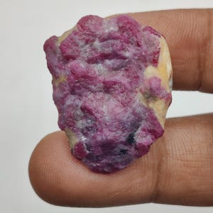 Puede incluir: Una gema de rubí en bruto, sin cortar, de un color rojo rosado intenso, salpicada de manchas blancas y amarillas. La piedra natural se sostiene entre los dedos, mostrando su superficie irregular y texturizada. Espécimen mineral.