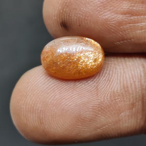 May include: Oval-shaped sunstone gemstone with a shimmering, golden-orange color. The stone has a polished surface and displays a sparkling effect. The gemstone is held between two fingers, showcasing its size and color.