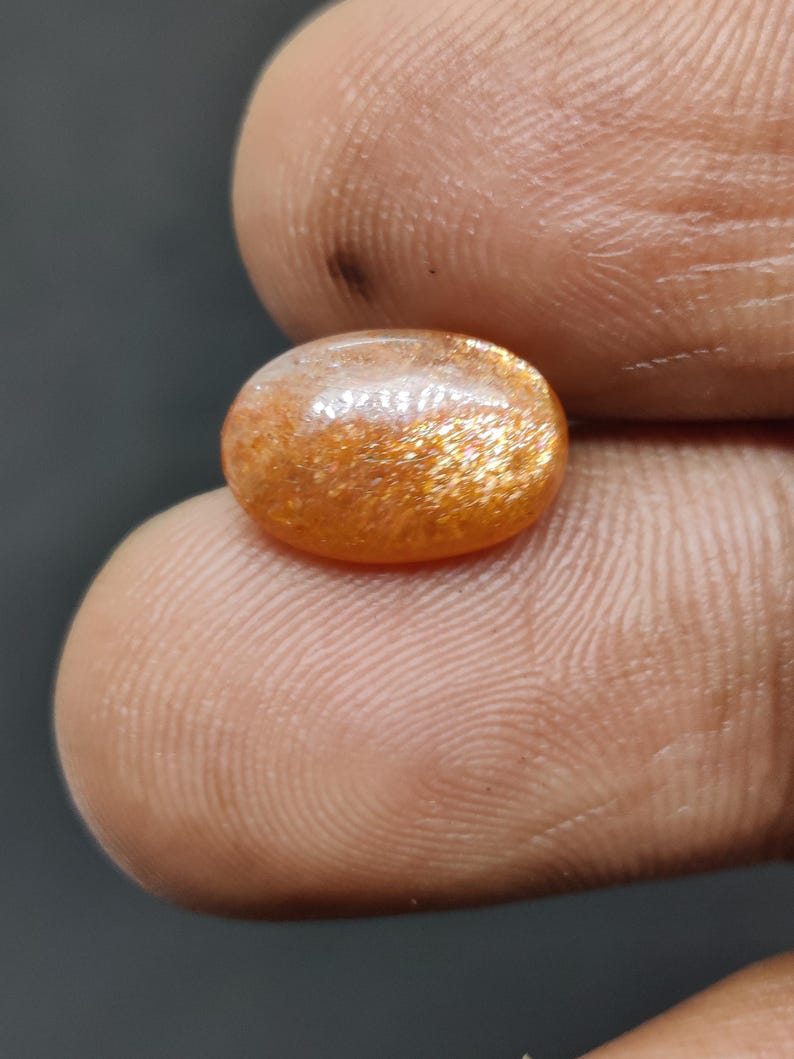 May include: Oval-shaped sunstone gemstone with a shimmering, golden-orange color. The stone has a polished surface and displays a sparkling effect. The gemstone is held between two fingers, showcasing its size and color.