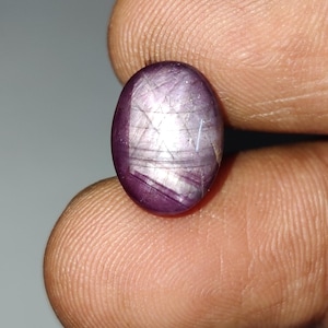 May include: An oval-shaped cabochon gemstone with a deep purplish-red hue. The surface displays a distinct six-rayed star effect, known as asterism, created by light reflecting off internal needle-like inclusions within the polished stone.