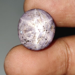 May include: A round, light purple gemstone with a star-like effect. The surface has a textured appearance with streaks and small dark inclusions. The gem is held between two fingers, showcasing its size and detail.