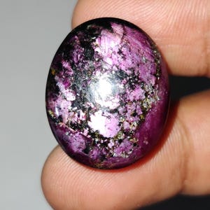 May include: An oval gemstone, predominantly magenta with black and gold inclusions. The polished surface reflects light, revealing a unique pattern. The gemstone is held in a hand, emphasizing its size and colour. The stone is likely a cabochon.