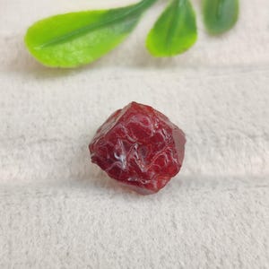 May include: A small, raw, deep red gemstone with a faceted, irregular shape. The stone displays a translucent quality with visible internal textures and a glossy surface finish, showcasing a rich, saturated crimson colour throughout its structure.