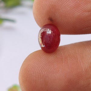 Puede incluir: Una gema pulida, de forma ovalada, de color rojo intenso. La gema se sostiene entre dos dedos, mostrando su superficie lisa y reflectante. El color de la piedra es un rojo rico y saturado, con una ligera translucidez.