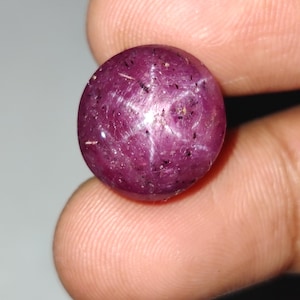 Può includere: Una gemma di rubino stellato di forma rotonda e taglio cabochon, di colore rosso magenta intenso. La pietra presenta un distinto effetto a stella a sei raggi, noto come asterismo, visibile sulla sua superficie lucida e traslucida.