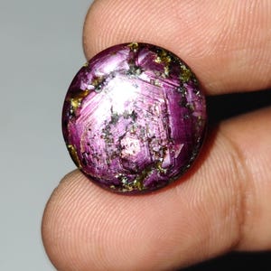 Puede incluir: Una gema redonda y pulida de color magenta intenso, que exhibe un patrón en forma de estrella. La gema tiene una superficie ligeramente irregular con motas de oro y negro. La apariencia general sugiere una calidad natural y sin refinar.