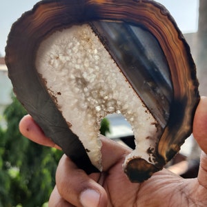 Puede incluir: Una geoda de ágata marrón y blanca con un agujero natural en el centro. La geoda tiene una superficie lisa y pulida y una variedad de colores marrones, blancos y negros.