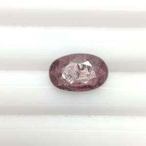 Puede incluir: Gema facetada de forma ovalada con un tono marrón rojizo intenso. La gema muestra reflejos internos y una superficie pulida. La piedra se encuentra sobre un fondo blanco con líneas paralelas, realzando su atractivo visual. Adecuada para la creación de joyas.