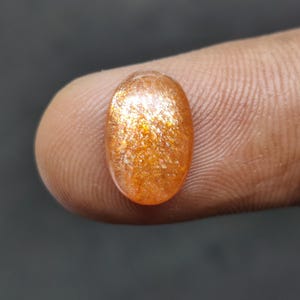May include: Oval-shaped, orange gemstone with a sparkling, glittery appearance. The gem is a warm, golden color with flecks of light throughout. The stone is held between two fingers against a dark background.