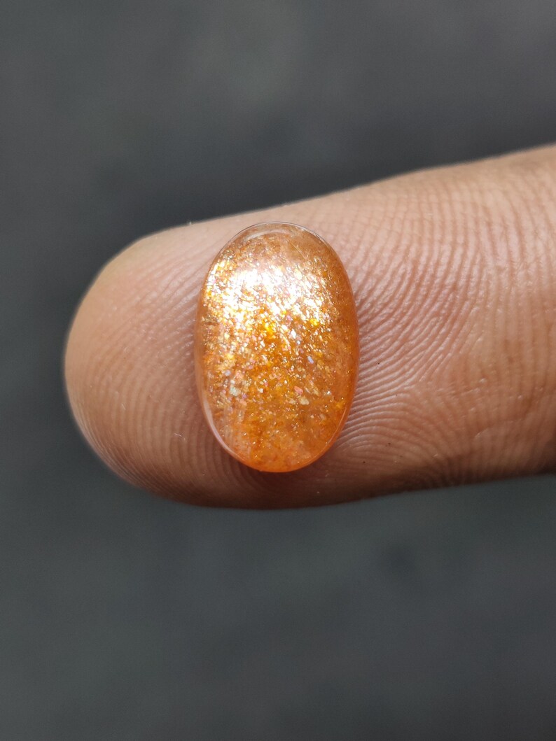 May include: Oval-shaped, orange gemstone with a sparkling, glittery appearance. The gem is a warm, golden color with flecks of light throughout. The stone is held between two fingers against a dark background.