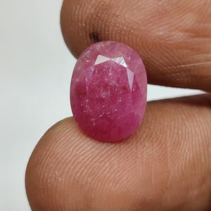 Könnte beinhalten: Ein ovaler, facettierter Edelstein mit einem tiefen rosa-roten Farbton. Der Stein hat eine polierte Oberfläche mit mehreren dreieckigen Facetten, die das Licht reflektieren und ein durchscheinendes Inneres mit natürlichen Einschlüssen im gesamten Kristall zeigen.