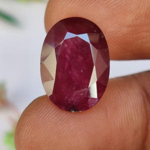 Könnte beinhalten: Ein oval geschliffener, facettierter Rubin-Edelstein. Der Edelstein hat eine tiefe, satte rote Farbe mit mehreren Facetten, die das Licht reflektieren. Der Rubin wird zwischen zwei Fingern gehalten, was seine Größe und Brillanz zeigt.