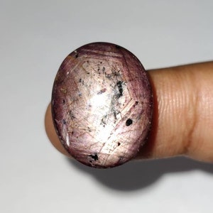 Puede incluir: Gema ovalada de color rosa púrpura. La superficie pulida de la piedra muestra inclusiones y pequeños puntos oscuros. La gema se sostiene entre los dedos, resaltando su forma. Esta gema probablemente está destinada a la joyería.