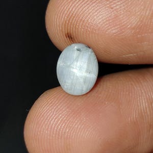 May include: An oval-shaped, pale blue gemstone with a subtle, shimmering effect. The stone has a smooth surface and appears to be held between two fingers against a dark background. The gemstone's color is a soft, muted blue-gray.
