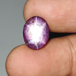 Pode incluir: Gema ovalada, vermelho-púrpura, com efeito de estrela. A gema tem uma superfície polida e um padrão de estrela proeminente visível no centro. A pedra é segurada entre os dedos, mostrando sua cor e forma.