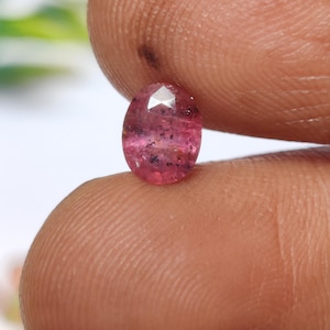 Puede incluir: Una gema rosa facetada de forma ovalada. La gema tiene una apariencia translúcida con inclusiones internas. La piedra se sostiene entre dos dedos, mostrando su tamaño y color. Las facetas de la gema capturan la luz, creando un efecto brillante.