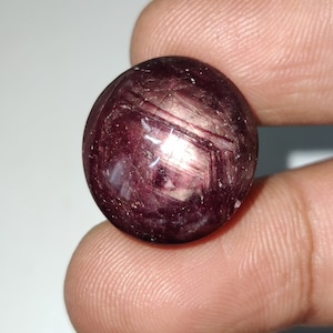 Könnte beinhalten: Ein runder, cabochon geschliffener, roter Edelstein mit einem sternförmigen Muster. Der Edelstein hat eine tiefrote Farbe mit einem leichten Schimmer.