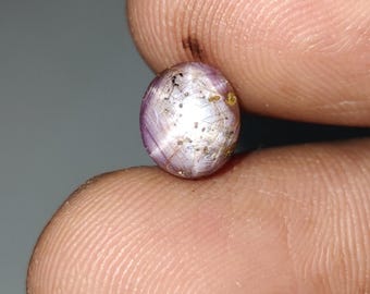 May include: A close-up of a star ruby gemstone with a six-rayed star. The ruby is a light purple color with a white star and gold inclusions.