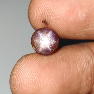May include: A round, cabochon-cut star ruby gemstone with a purplish-red hue. The stone displays a distinct six-rayed star effect, known as asterism, visible across its polished, translucent surface, which features natural internal inclusions.