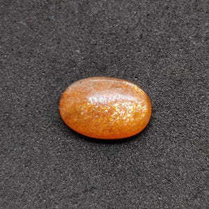May include: Oval-shaped sunstone gemstone with a shimmering, orange and gold appearance. The polished stone has a smooth surface and is set against a dark, textured background. This gemstone is suitable for jewelry making.