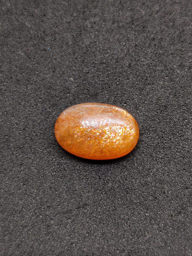 May include: Oval-shaped sunstone gemstone with a shimmering, orange and gold appearance. The polished stone has a smooth surface and is set against a dark, textured background. This gemstone is suitable for jewelry making.