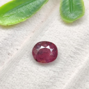 May include: An oval-shaped, faceted gemstone in a deep magenta-pink hue. The stone features a polished surface with visible dark, natural inclusions scattered throughout its interior, creating a unique pattern within the translucent material.
