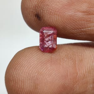 May include: A rectangular, faceted gemstone with a vibrant pinkish-red hue. The stone displays internal inclusions and a clear, polished surface, showcasing a classic emerald-style cut with sharp corners and multiple reflective facets throughout.