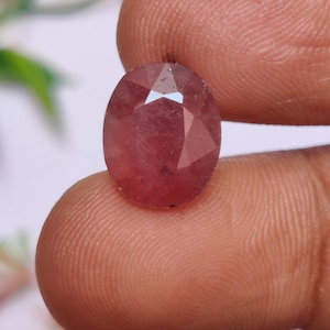 Puede incluir: Una gema facetada de forma ovalada con un tono rosa rojizo. La gema se sostiene entre los dedos, mostrando sus superficies pulidas y claridad interna. Las facetas capturan la luz, creando un efecto brillante. Esta gema es adecuada para joyería.